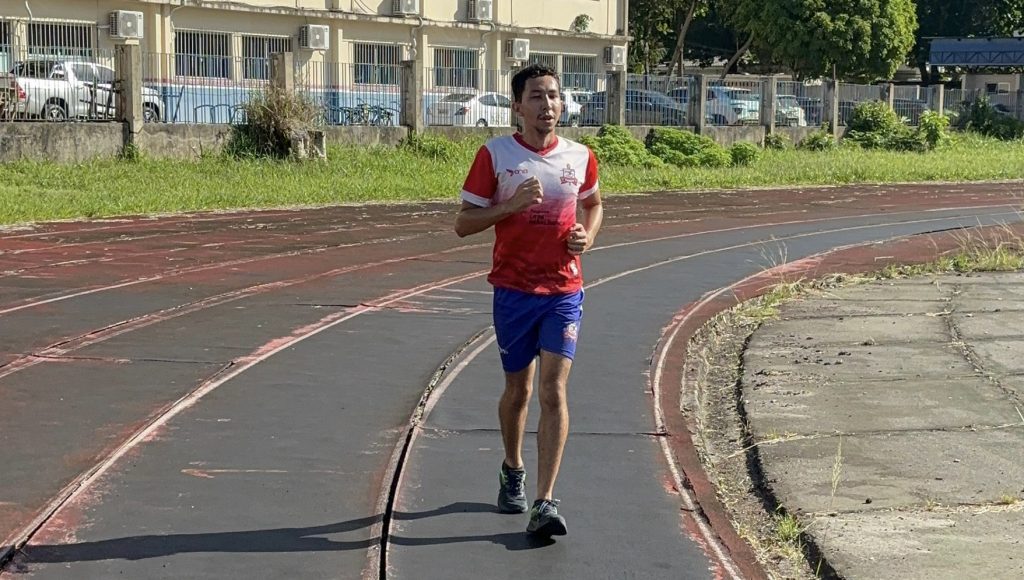 Fotografia registra um praticante de atletismo. O homem veste uma camisa com brasão da UFPA, short com brasão da UFPA, e usa tênis. Ele se movimento em uma longa pista de atletismo.